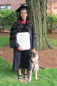 Photo de Haben GIRMA en tenu de remise de diplome américain (grande robe noir semble à celles des avocats en France), tenant son diplôme à la main. Son chien guide se tient assis à côté d'elle. 