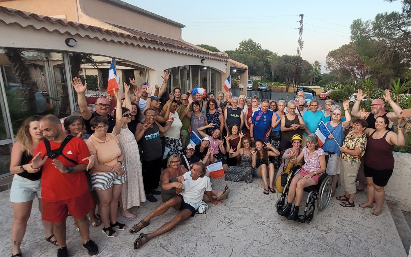 Photo de la session rencontre à Frejus en Juin 2026 représentant un groupe très nombreux de personnes souriantes rassemblées en extérieur devant un bâtiment clair avec toit en tuiles. Les personnes sont de tous âges, hommes et femmes, avec différentes morphologies et couleurs de peau claires à moyennes. La majorité des gens ont les bras levés ou font des signes de la main, l’ambiance est festive et détendue. Plusieurs drapeaux français sont tenus par des personnes réparties dans le groupe, et un petit drapeau bleu‑blanc‑rouge est aussi planté près du centre.  
Les vêtements sont d’été : shorts, robes légères, T‑shirts, débardeurs, sandales et baskets, avec quelques inscriptions visibles comme “ITALIA” sur un débardeur vert et “SPORTMAX” sur un T‑shirt sombre. Une femme à droite est assise dans un fauteuil roulant, en short rose et chemisier fleuri, à côté d’autres personnes assises par terre ou accroupies qui rient et posent. Deux personnes sont allongées au sol au premier plan, un homme en T‑shirt blanc tenant une chaussure et une femme en robe courte kaki, tous deux en train de plaisanter avec le groupe.  
À l’arrière‑plan, la façade vitrée du bâtiment reflète l’intérieur, et plus loin se trouvent quelques arbres, une route et des voitures garées, sous un ciel clair de fin de journée.